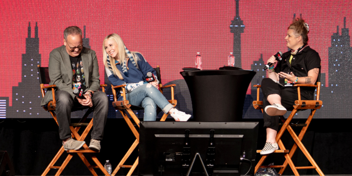 Dee Bradley Baker, Ashley Eckstein, and moderator Kristin Finger sit in front of a black and red Toronto Skyline backdrop. Dee Bradley Baker looks down in laughter, as Ashley Eckstein leans into him smiling.