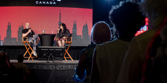 Christopher Sabat and moderator Jaclyn Thomas sit on stage in front of a red and black Toronto skyline. in the foreground, fans are waiting in line to ask questions at the microphone.