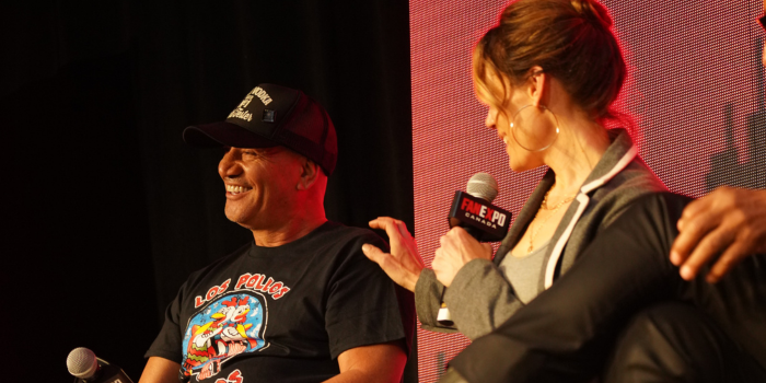 Emily Swallow grabs Temuera Morrison's shoulder as she tells a story. Temuera smiles toward the audience. They are holding FAN EXPO Canada microphones and sitting in front of a red and black LED screen.