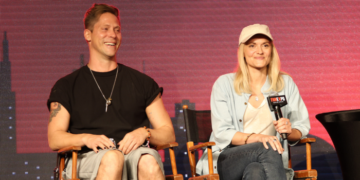 Neil Newbon and Patricia Summersett take the stage together. They are seated in director's chairs in front of a black and red image of the Toronto skyline on the LED screens behind them. Both are holding the FAN EXPO Canada branded microphones.