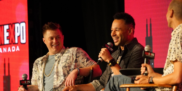 Left to right: Lucas Grabeel, Corbin Bleu, moderator Trevor Campbell. Lucas leans forward, smiling at Corbin as Corbin answers a fan question with a smile on his face as well.