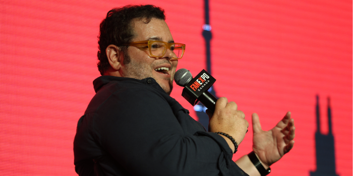 Josh Gad smiles as he speaks. He is seated in front of a red and black Toronto skyline LED background, holding a black FAN EXPO Canada branded microphone. 