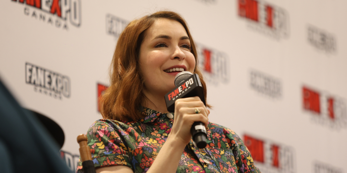 Felicia Day sits in front of the white FAN EXPO Canada backdrop, holding a FAN EXPO Canada microphone to her face. She smiles as she speaks.