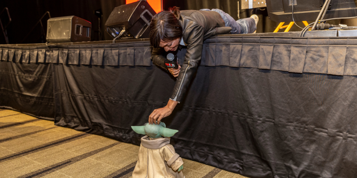 Rosario Dawson lays on the main stage to greet a small animatronic Grogu that has been piloted to meet her. She pets him on his liitle head.
