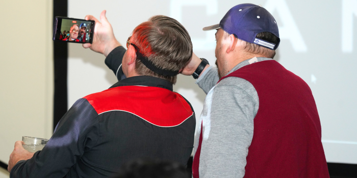 Bubbles holds the phone as he and a fan pose for a selfie, their backs to the screen. From over Bubbles' shoulder, you see the phone screen of the selfie being taken, where both parties are smiling