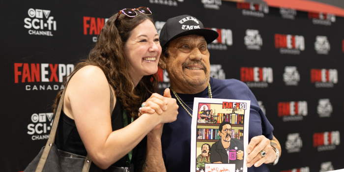 Danny Trejo holds up a piece of art brought to be signed by a fan. He and the fan pose together, holding hands as they smile for the camera.
