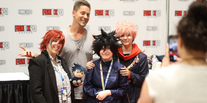 Neil Newbon poses with three fans dressed as various anime characters, one holds a plush of an additional character. Neil holds his arms behind them as if to present the group. A volunteer takes the photo on a smartphone 