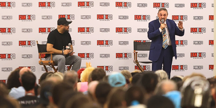 John Rhys-Davies addresses a very full room of fans. He holds the microphone to his face, while the moderator watches and listens intently to him.