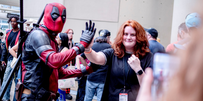 a fan dressed as Deadpool poses, high fiving a fan who is not in cosplay. In the foreground, a person takes a photo of the pair.