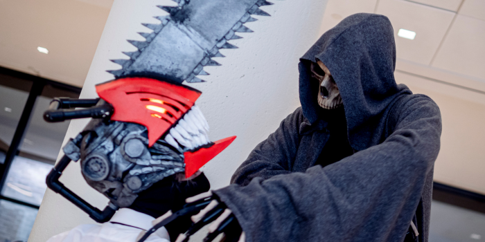 A cosplayer dressed as the Grim Reaper (on stilts to add height) poses, holding the shoulder of a cosplayer dressed as Denji from Chainsaw Man. 