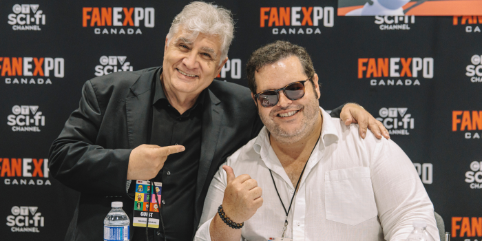 Maurice Lamarche joins Josh Gad at his table in the celebrity autograph area. The duo pose in front on the black FAN EXPO Canada backdrop, pointing at each other and smiling at the camera.