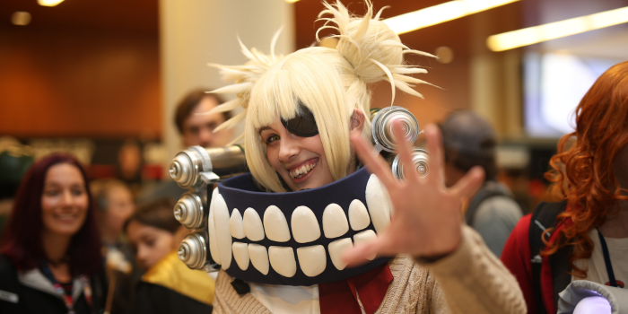 A cosplayer dressed as Himiko Toga smiles and reaches toward the camera. They have blonde hair in messy space buns, wearing a beige cardigan and red ascot, they have a collar around their neck that matches the vampire-esqe fangs they're wearing.