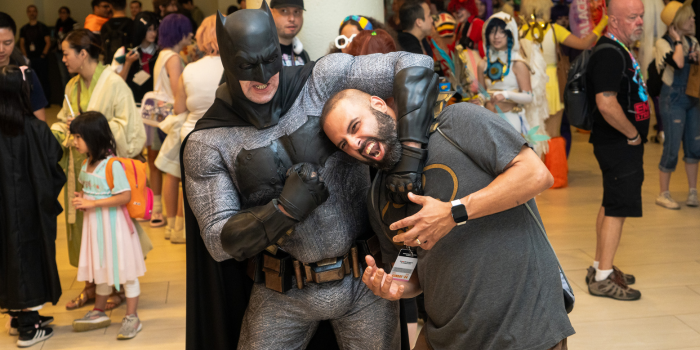 A Batman cosplayer holds a batman fan (dressed in a batman tshirt) in a headlock, Batman grits his teeth, as the other makes a surprised expression