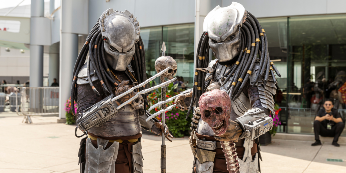 A pair of cosplayers stand outside, dressed as Predators from the movie Predator. One, holds a stake that brandishes a skull, the other, a bloodied skull still attached to a spine.