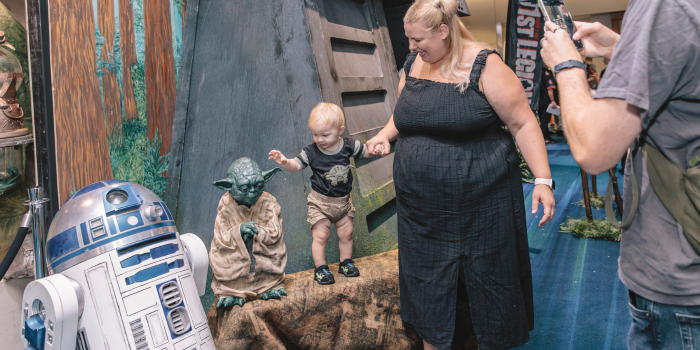 A mother poses with her child as the father takes a photo. The baby is stood on a bench next to a plastic Yoda, and an R2-D2.