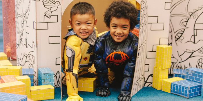Two kids, dressed as Bumblebee (Transformers) and Miles Morales (Into the Spider-verse) pop out of the door of a cardboard castle, surrounded by colourful cardboard bricks.