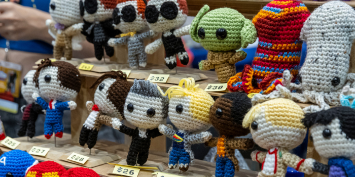 Crocheted characters lined up on a wooden platform. On the top line, the characters from Good Omens, Yoda, and Squids. One the bottom, all 13 of the doctors from Doctor Who.
