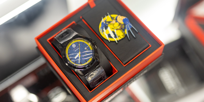 A watch in the box. On the left, the camera focuses on a gun metal grey watch with a yellow and blue face with white slash marks. Out of focus to the right is Wolverine, hanging out of the X-Men logo.