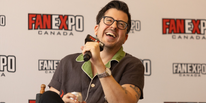 Christopher Mintz-Plasse sits in front of a white FAN EXPO Canada backdrop, he holds a silver can in his left hand and the microphone in his right. His head is cocked to the right as he smiles and laughs toward the audience.