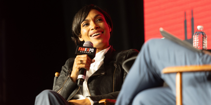 Rosario Dawson sits in a director's chair, holding a FAN EXPO Canada branded microphone. She smiles as she answers her moderator's question.