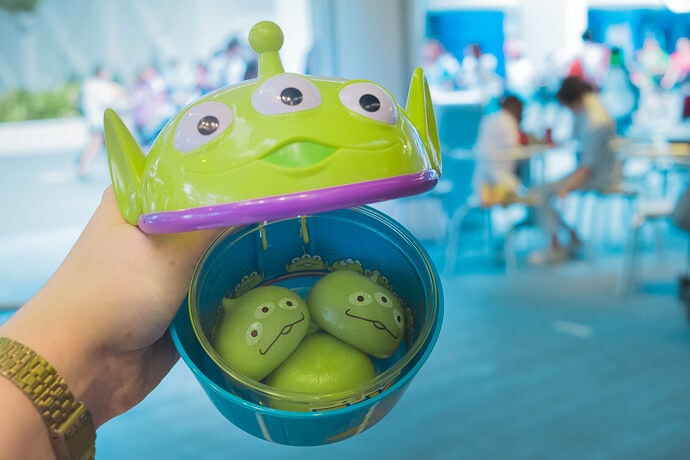 three little green men mochi in a little green men cup, inside of a little green man shaped cup. A hand holds the outer cup from the left side with a gold watch band. The background is a light blue, with diners at tables in the background