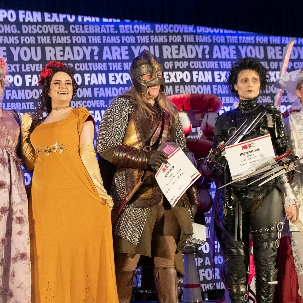 A group shot of the craftmanship cup winners. From the left, (cut off slightly) Bridgerton inspired Rapunzel and Belle, Battle Eowyn, and Edward Scissor Hands. They are holding their winners certificates. In the background, is the FAN EXPO background that repeats FAN EXPO and other Phrases.