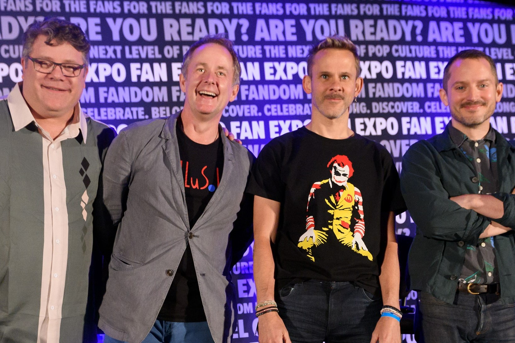 Group shot of Sean Astin (in a button up shirt with a white line through the middle, gray on either side and a black, white and gray diamond pattern on the right side of his shirt), Billy Boyd (gray jacket with a black shirt underneath with red words), Dominic Monaghan (black shirt with a Heath Ledger's Joker as Ronald McDonald, jeans, and a lot of wrist bands) and Elijah Wood (crossed armed, in a dark green jacket, dark gray Hawaiian shirt and dark Jeans). They are smiling at the audience. In the background, is the FAN EXPO background that repeats FAN EXPO and phrases.