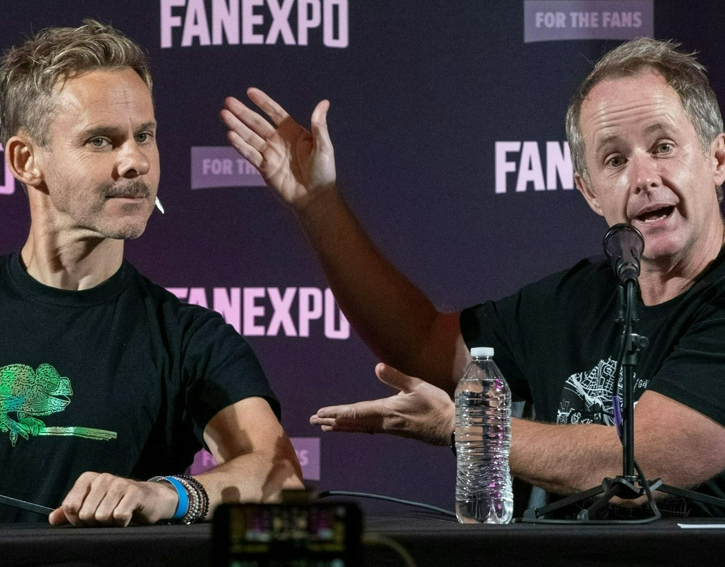 Dominic Monaghan and Billy Boyd hosting their Friendship Onion Podcast. Billy is gesturing to Dominic like he is presenting him to the audience. Dominic is posing proudly as he wears a black shirt with a chameleon on it. In the background, the words FAN EXPO are repeated.