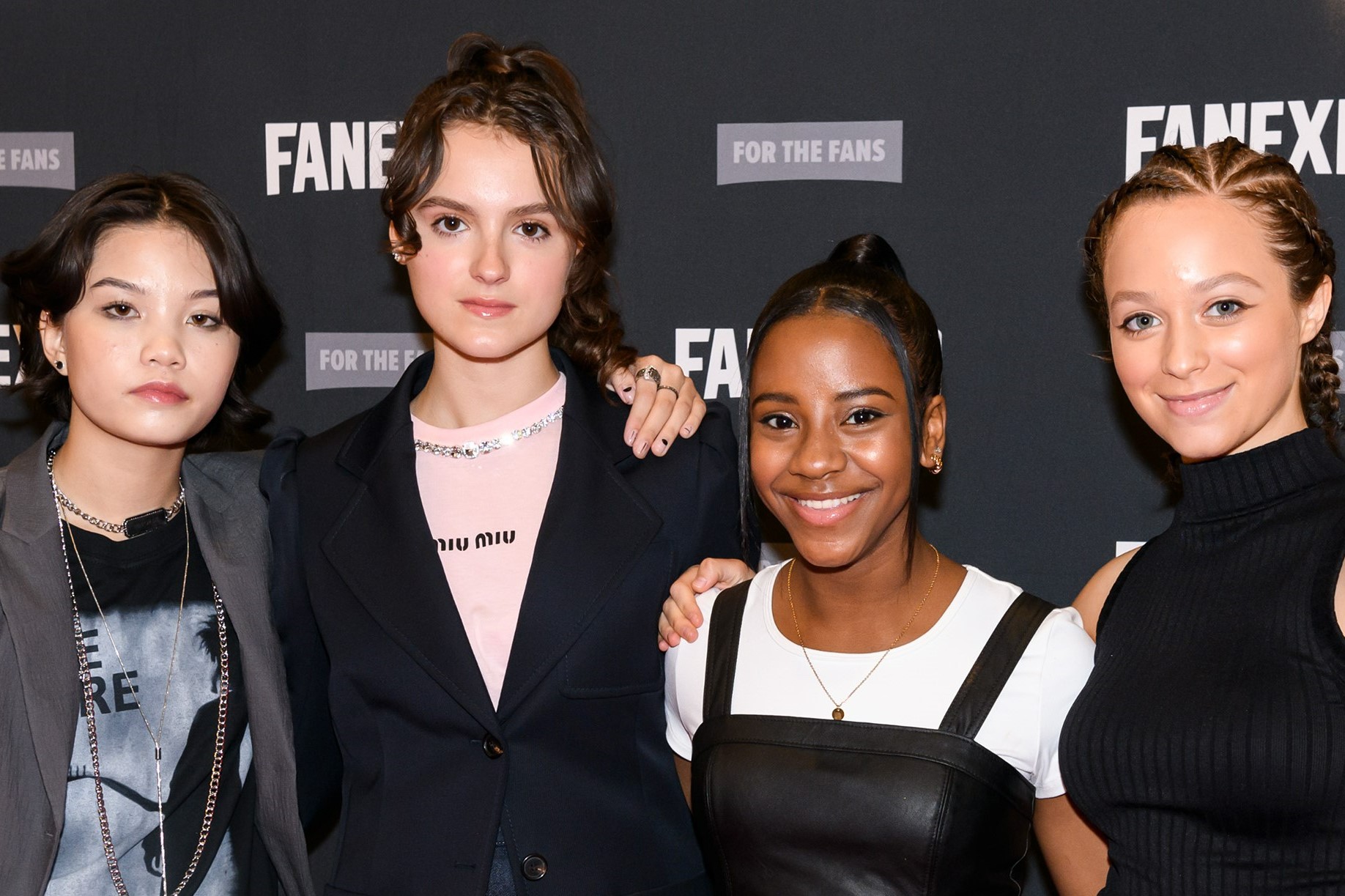 Group shot of the cast from Paper Girls:  Camryn Jones, Riley Lai Nelet, Sofia Rosinsky, and Fina Strazza. They are all wearing dark clothes and the two on the left are posing with a straight face while the two on the right are smiling. They are smiling at the audience. In the background, is the FAN EXPO background that repeats FAN EXPO.
