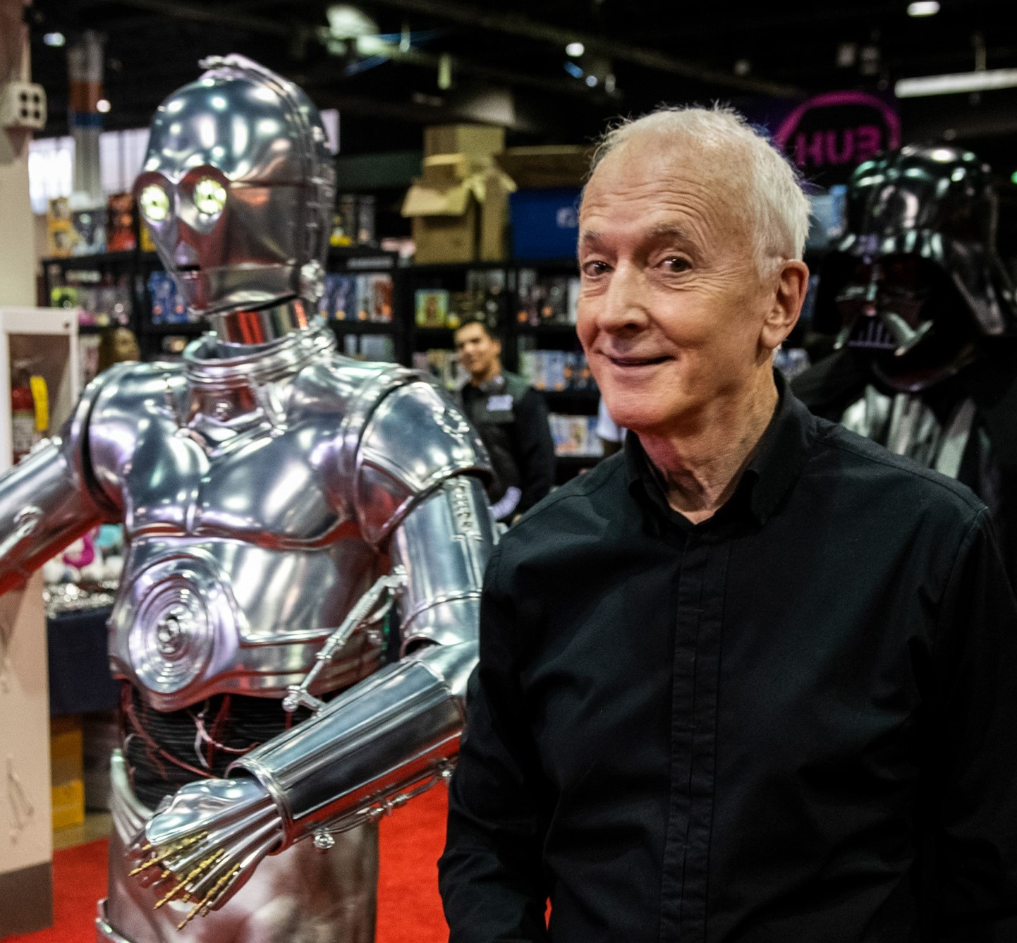 Anthony Daniels smiling as he walks with a silver C-3PO on the red carpet.