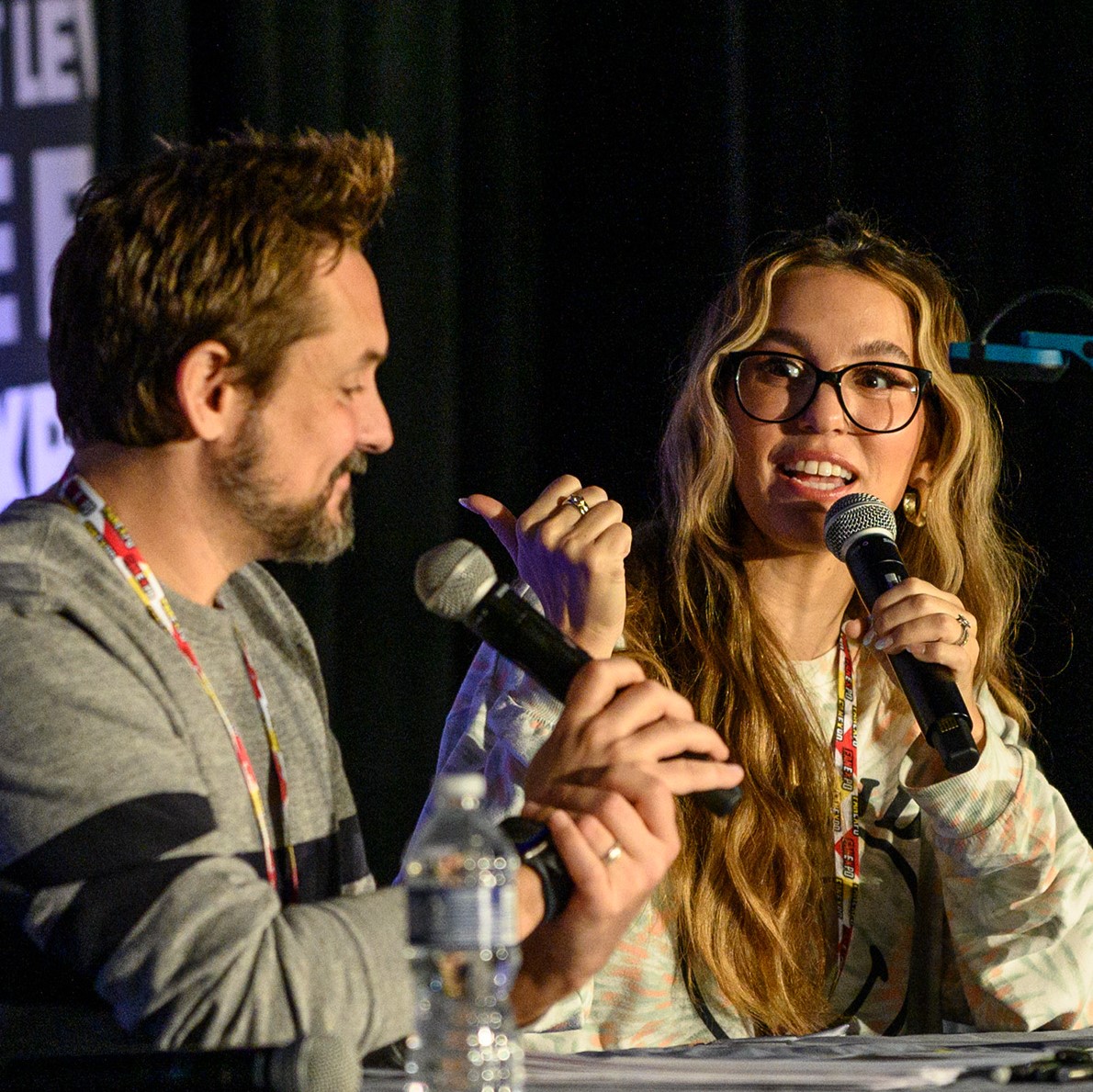 This is an angled shot of Will Friedle and Christy Carlson Ramano as they hold their mics and talk to each other. Will is wearing a gray long sleeve with the white/yellow/red FAN EXPO lanyard. Christy is wearing a light pastelish green and orange long sleeve shirt with a smiley face on it and she is wearing black glasses.