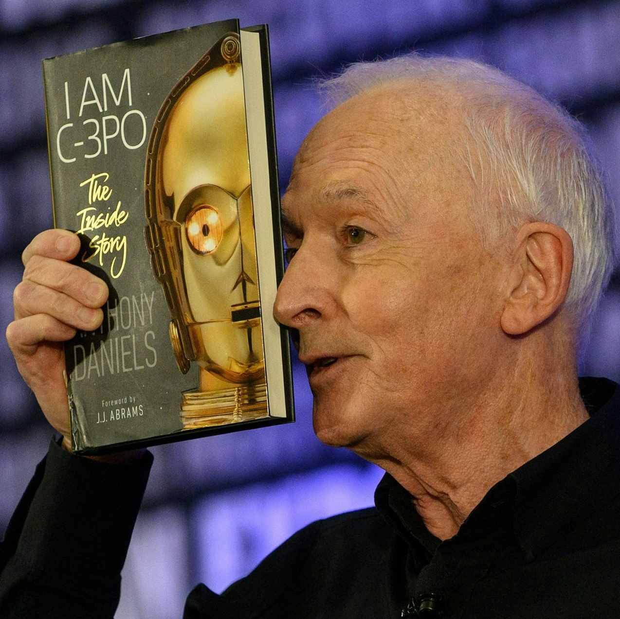 A head shot of Anthony Daniels holding his book "I AM C-3PO: The Inside Story by Anthony Daniels, Featuring JJ Abrams." with half of an image of C-3PO. He is holding the book to his face. He has short white hair and is wearing a black long sleeve shirt. In the background, is the FAN EXPO background that repeats a variety of phrases.