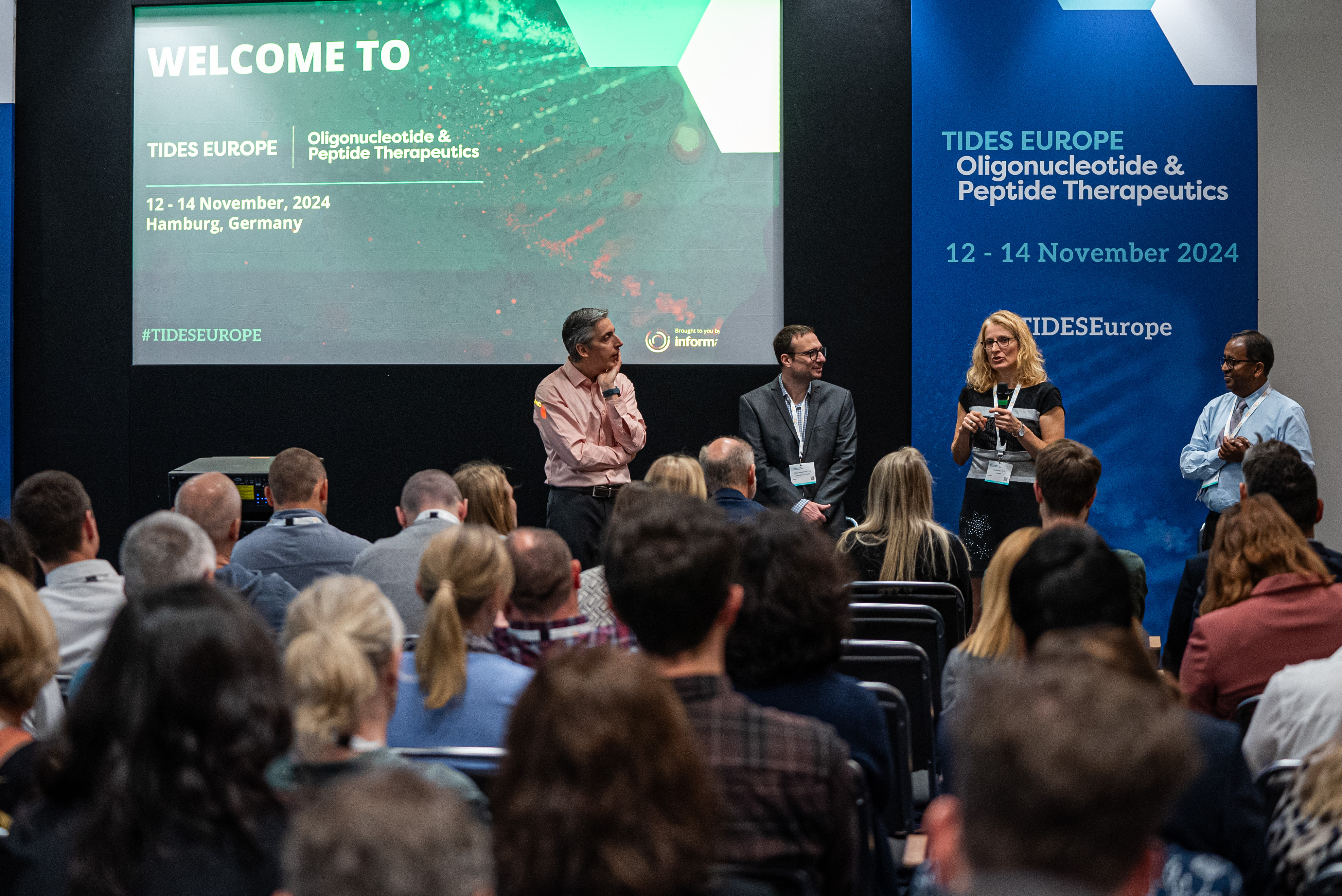 Four speakers presenting in front of an audience in a conference room with TIDES Europe posters and screen in the background