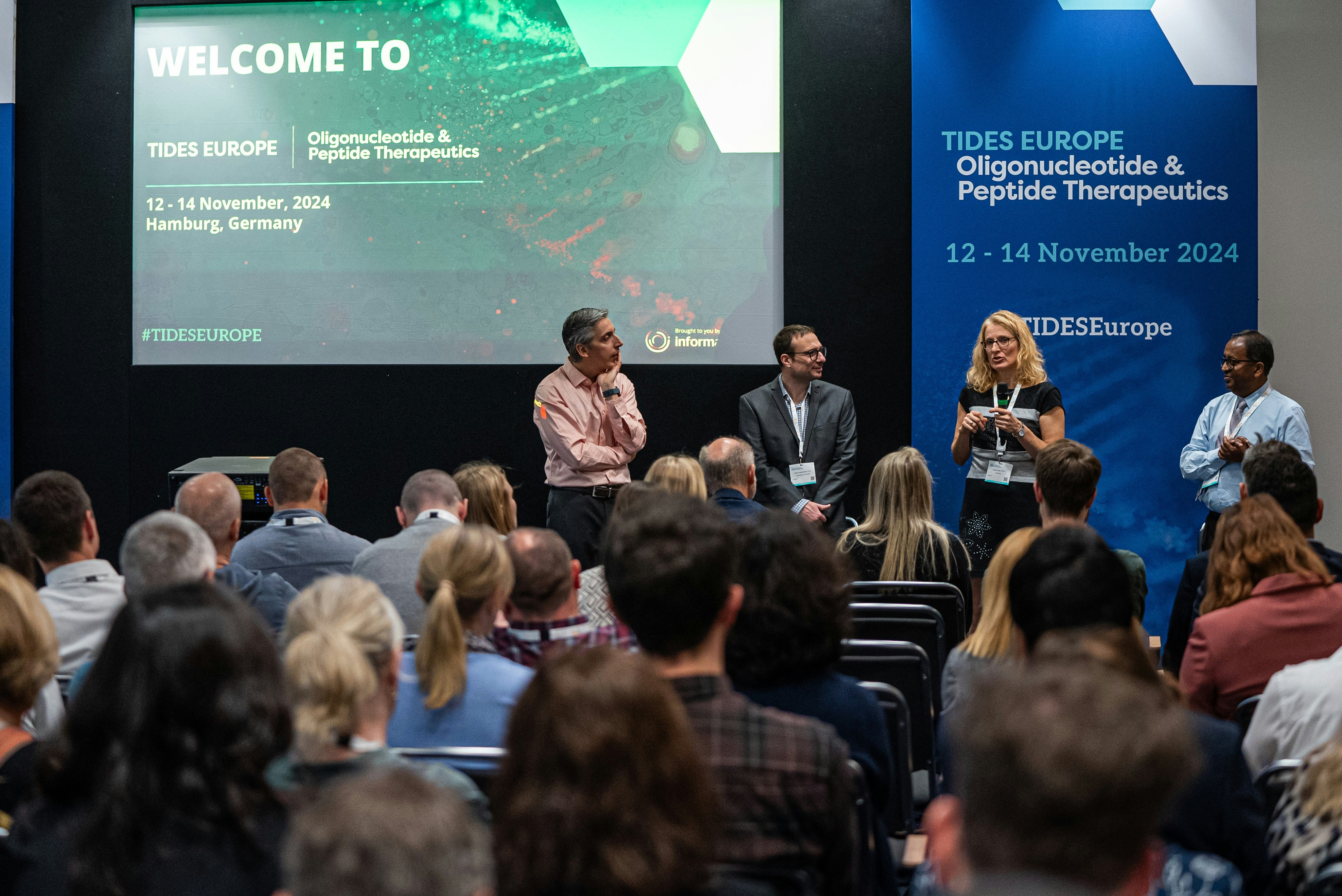 Four speakers presenting in front of an audience in a conference room with TIDES Europe posters and screen in the background