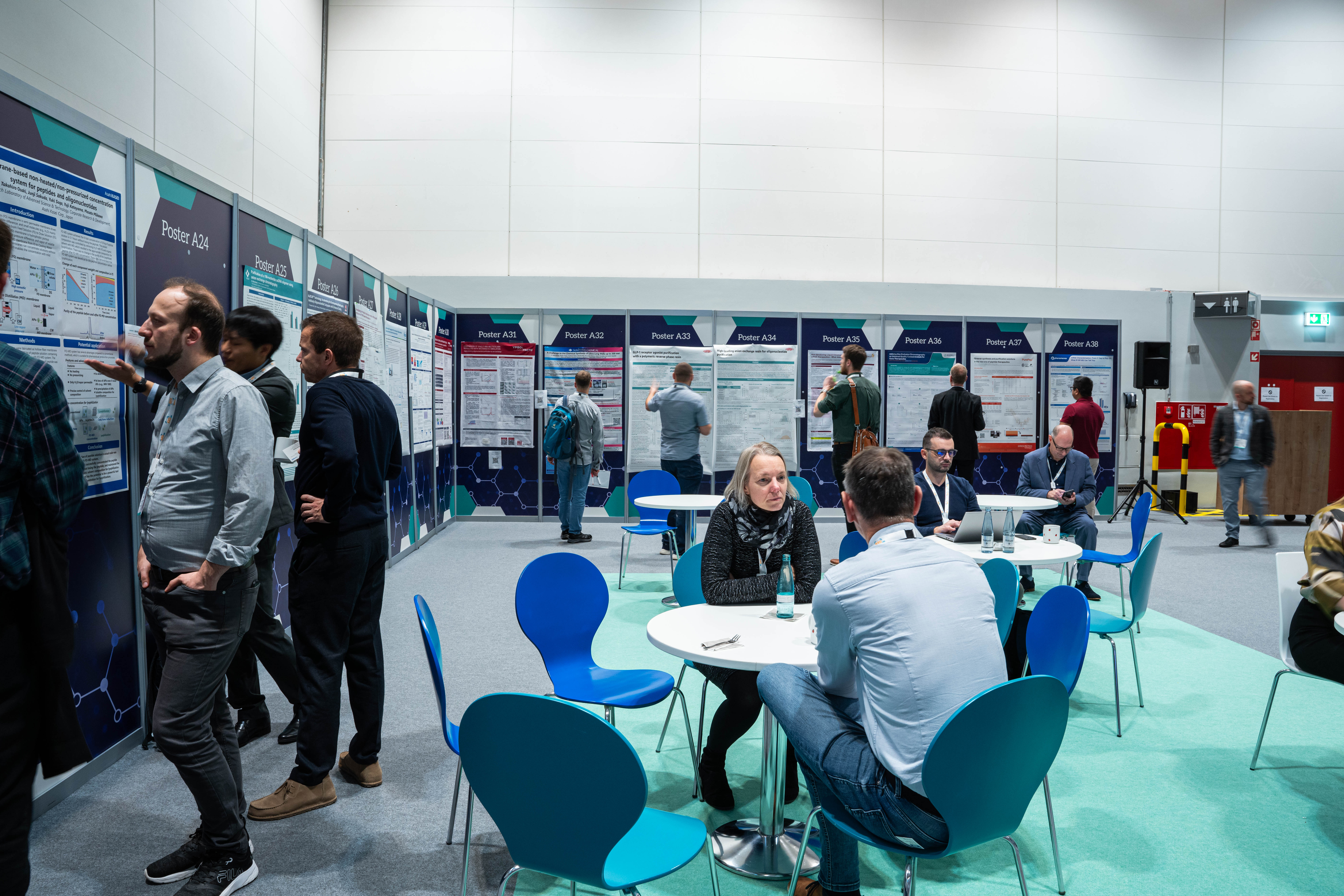 Two people are in discussion sat by table and chairs whilst others browse a poster wall.