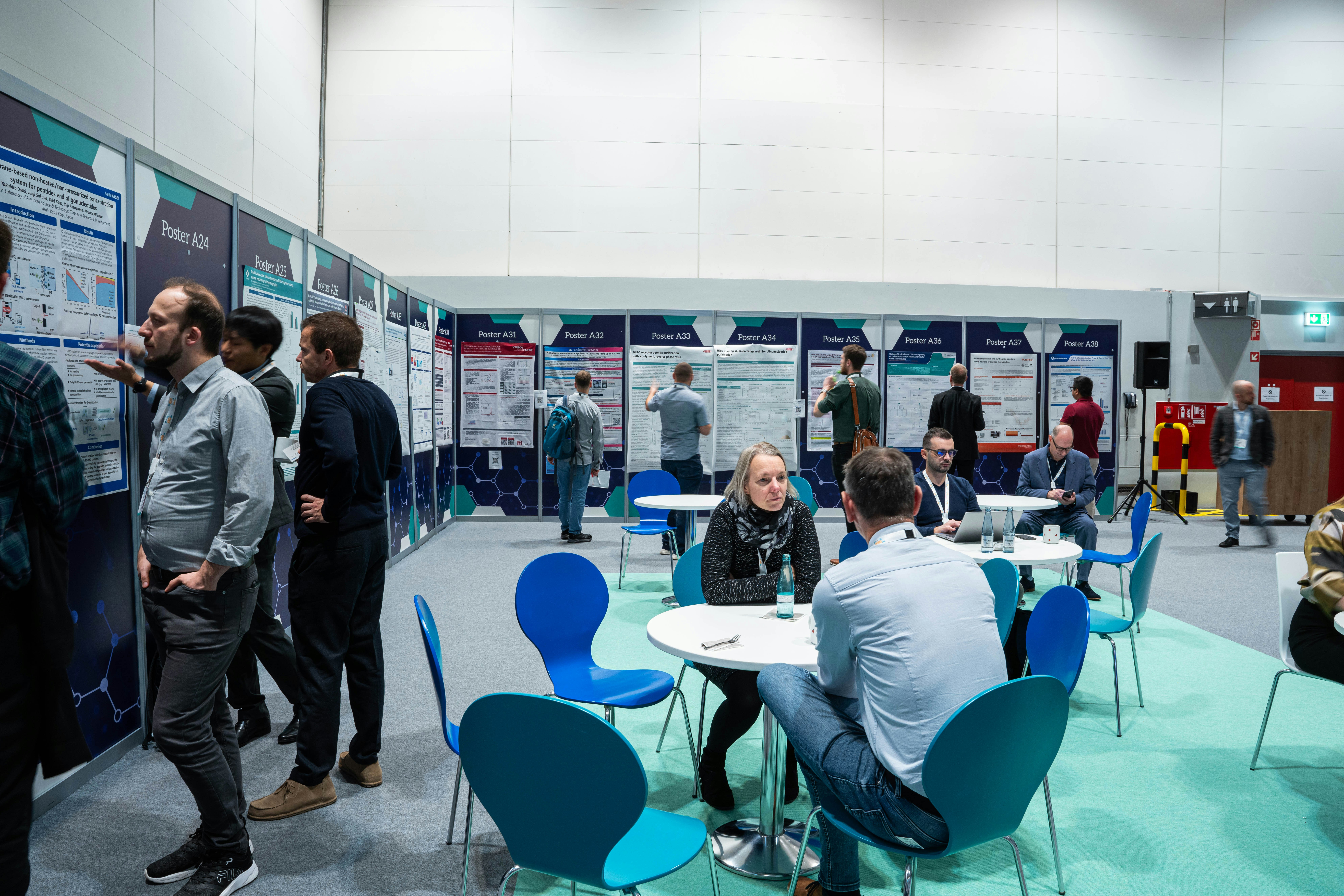 Two people are in discussion sat by table and chairs whilst others browse a poster wall.