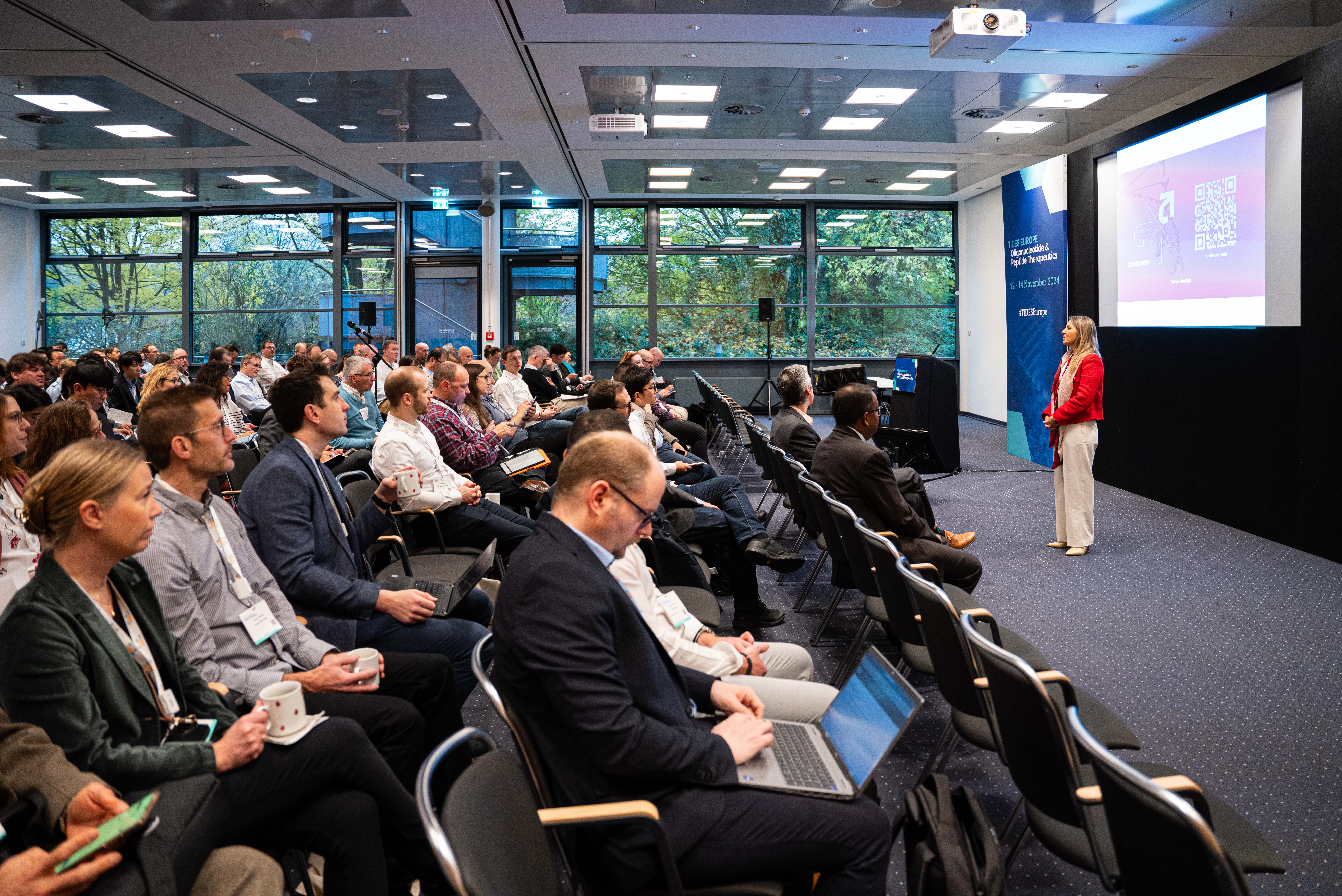 Attendees watching a female speaker in a red blazer talk at the front of the room