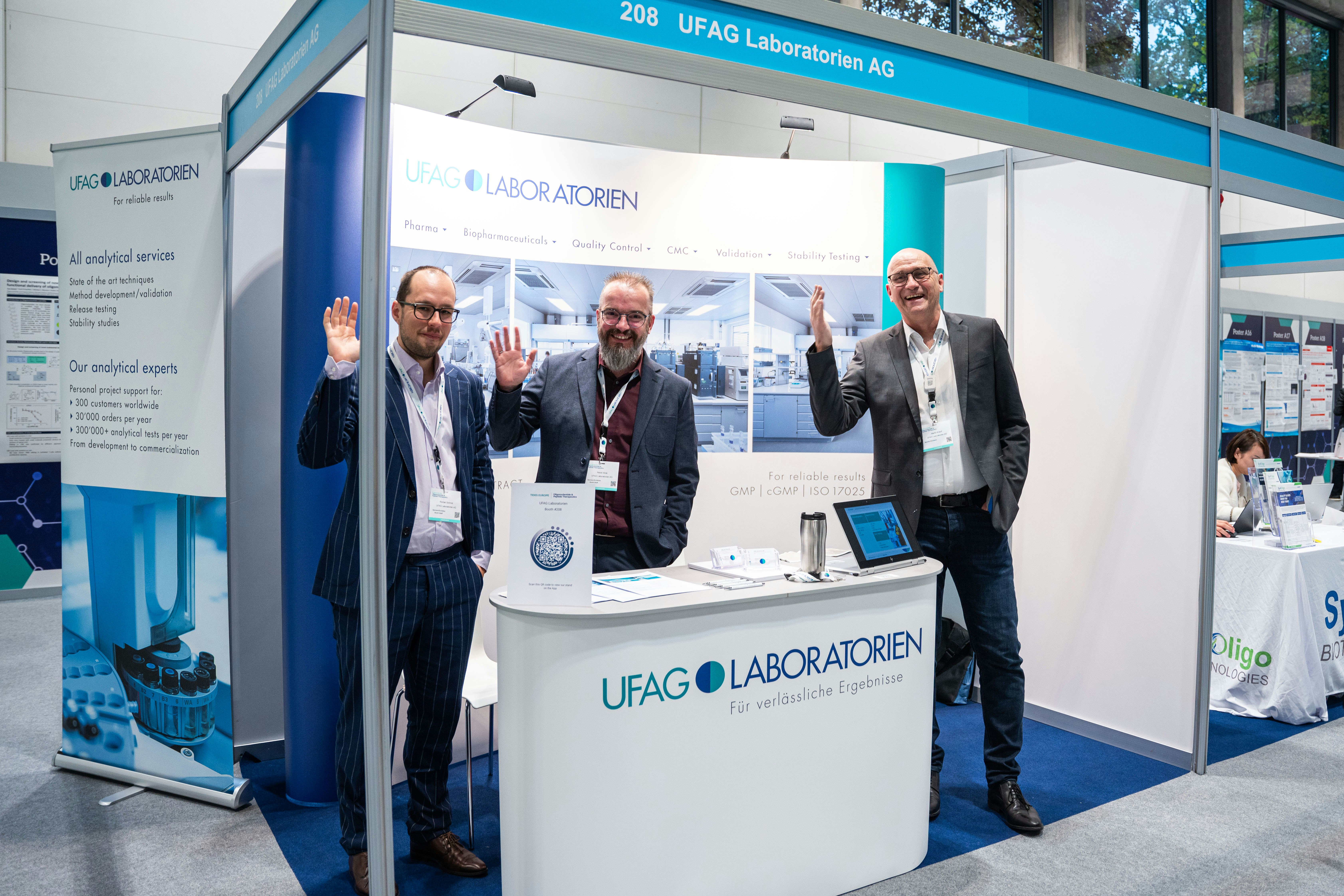 Three men in suits wave for the camera by a UFAB Laboratorien AG stand.