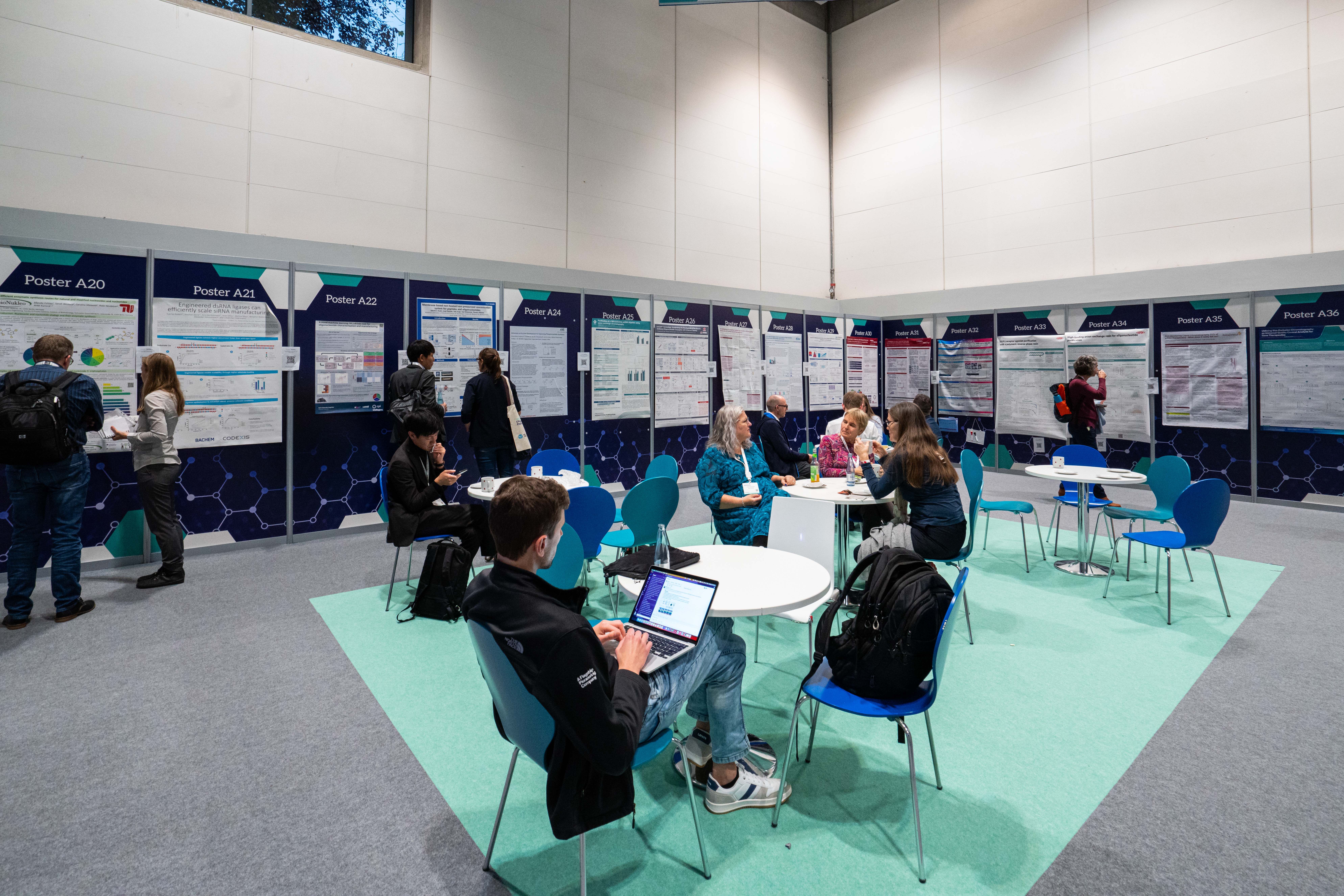 Tables and chairs set up by a wall of posters as some people sit and talk or sit on their laptop.