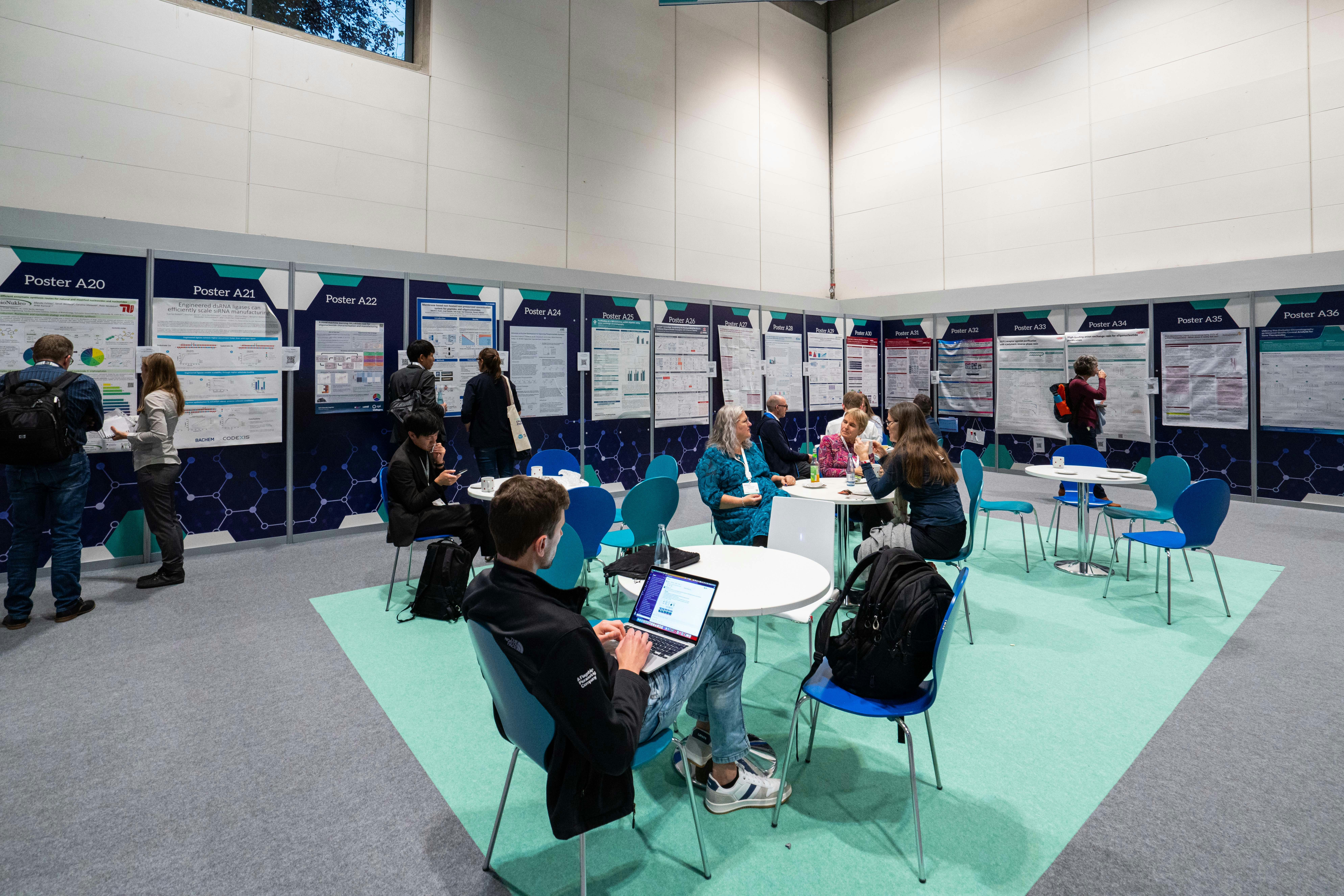 Tables and chairs set up by a wall of posters as some people sit and talk or sit on their laptop.