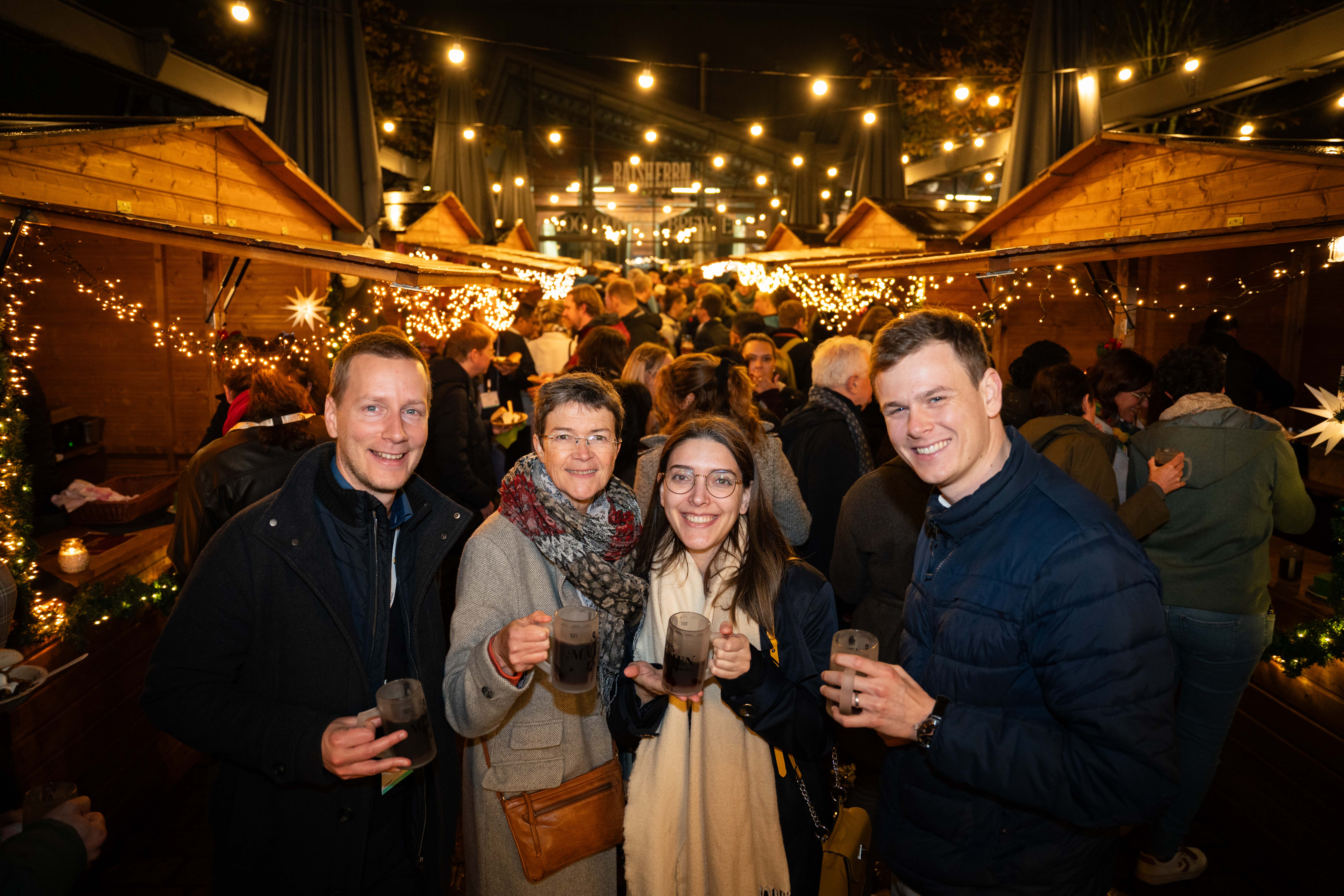 Four people smile for the camera with drinks in hand amidst a crowd of people outside at night with fairy lights