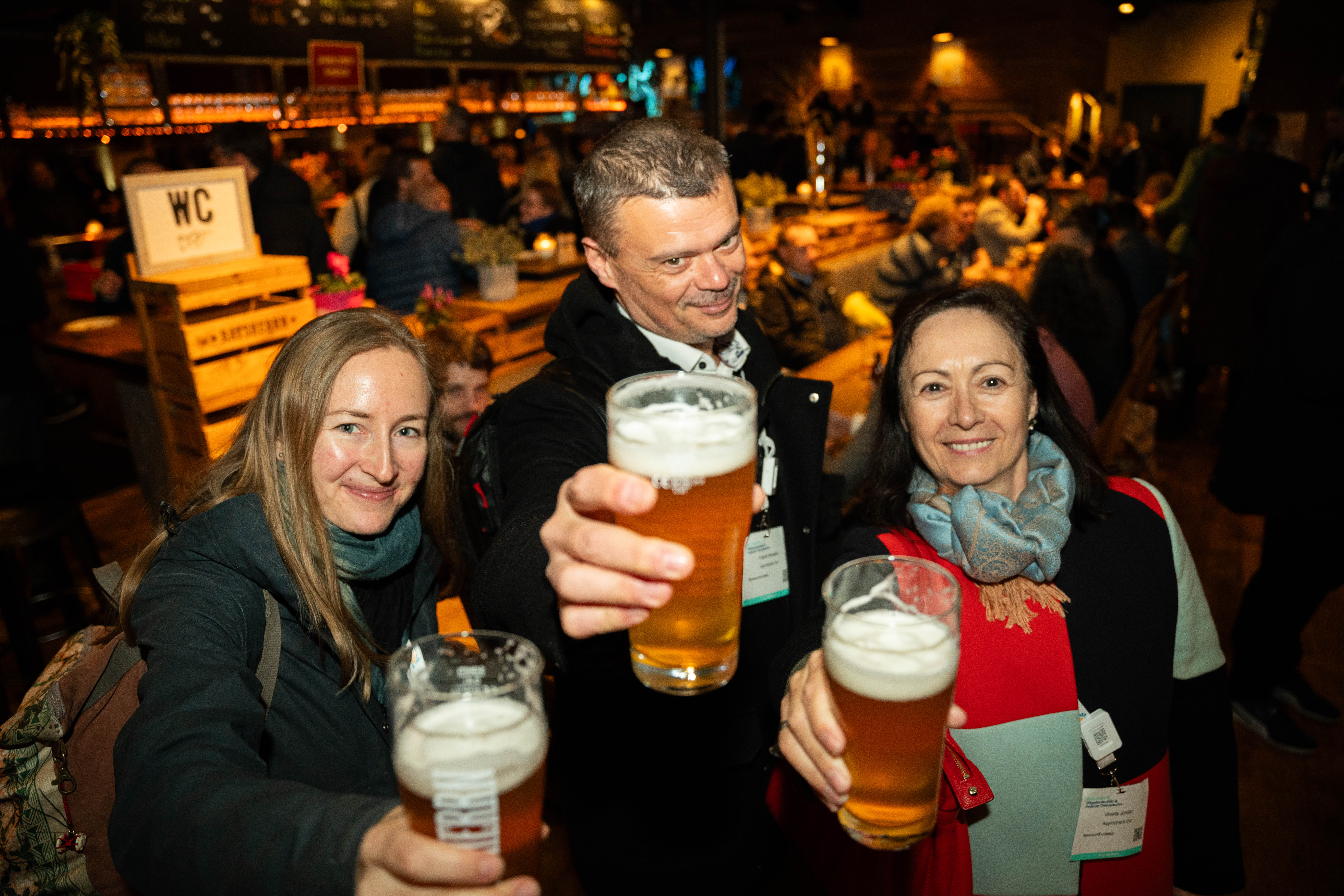 Two women and a man smile while holding their beers up for the camera