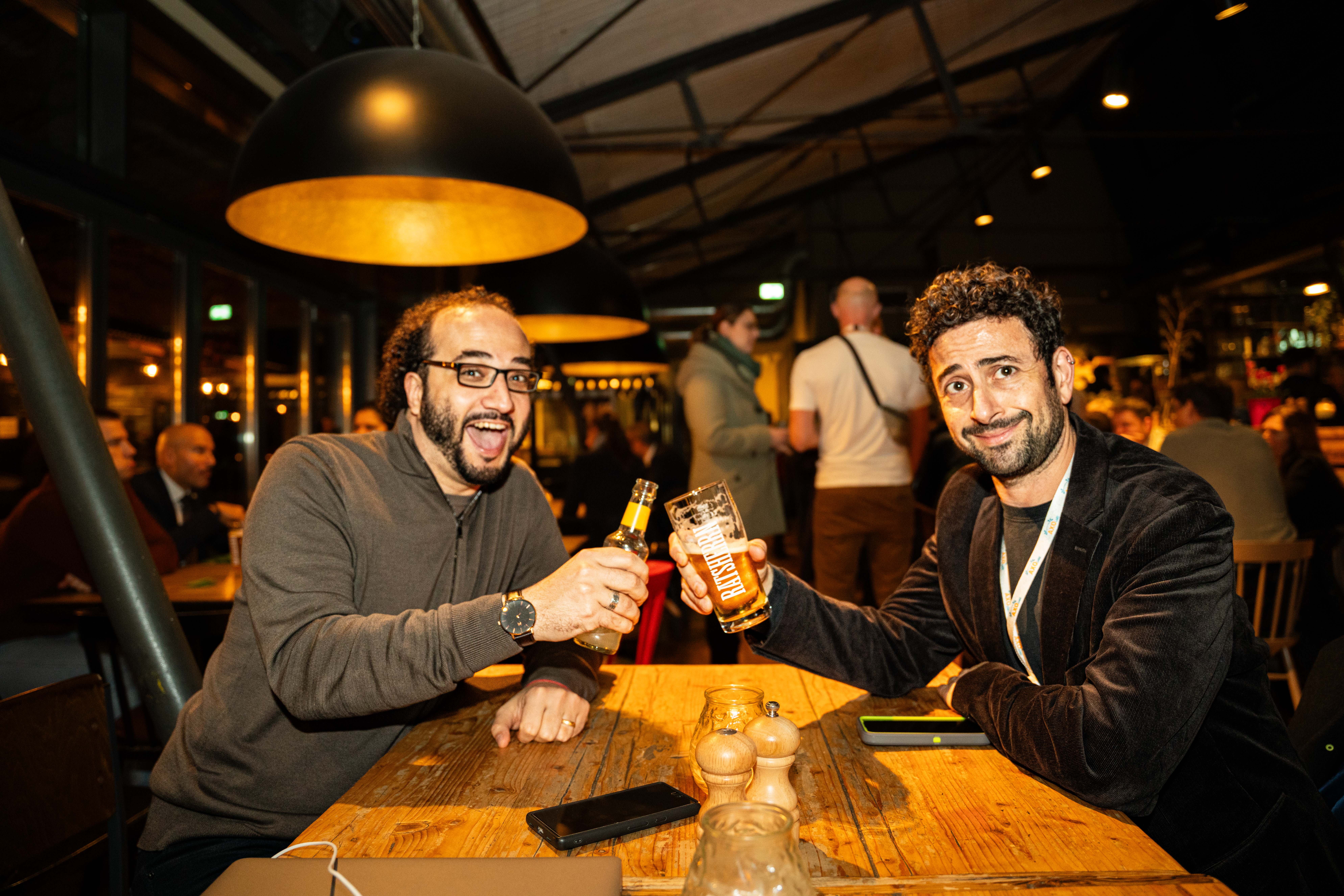 Two men cheers with their drinks and smile for the camera at a bar.