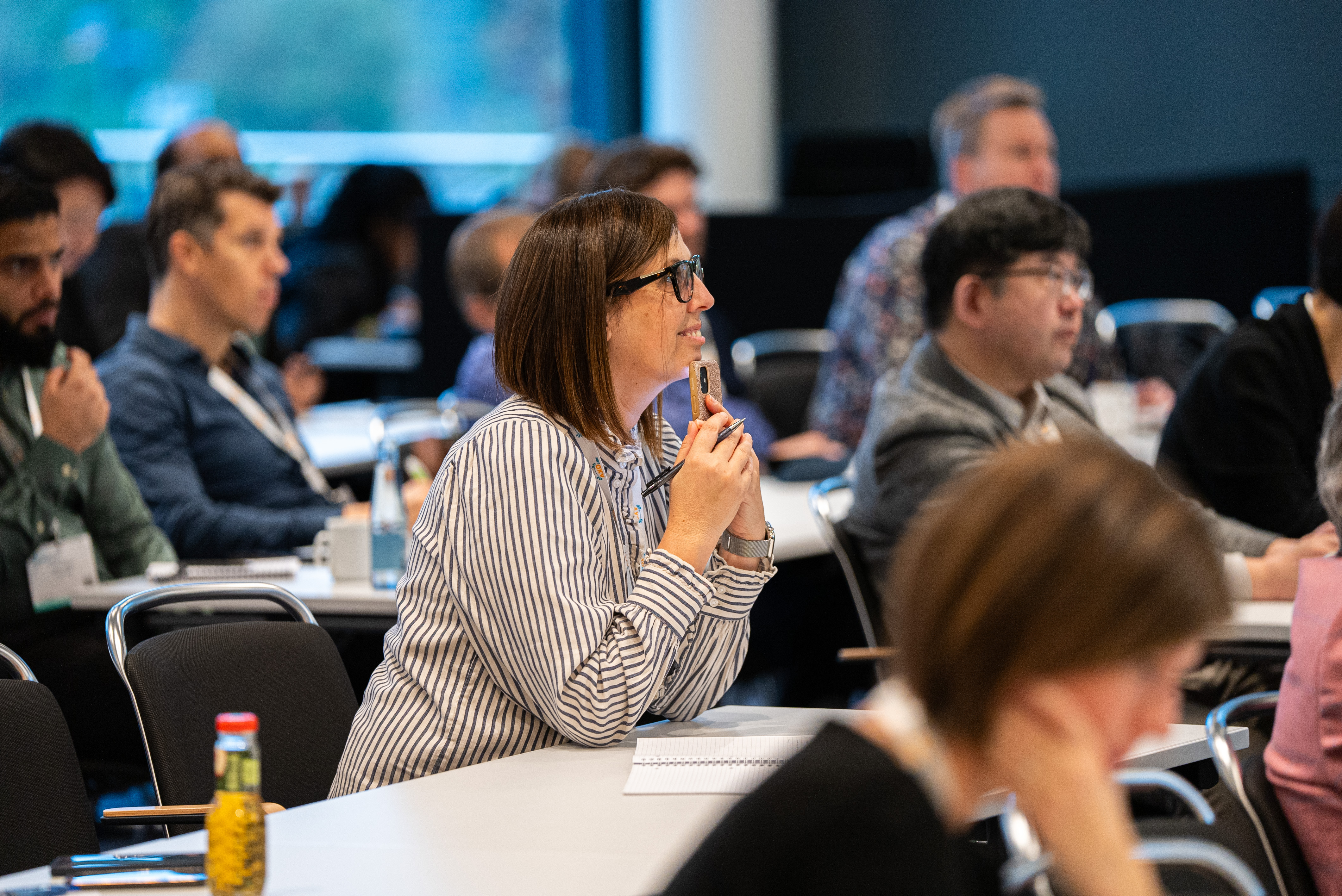 One woman in a stripe shirt smiles whilst sitting in a conference room, other attendees also feature sat in the back