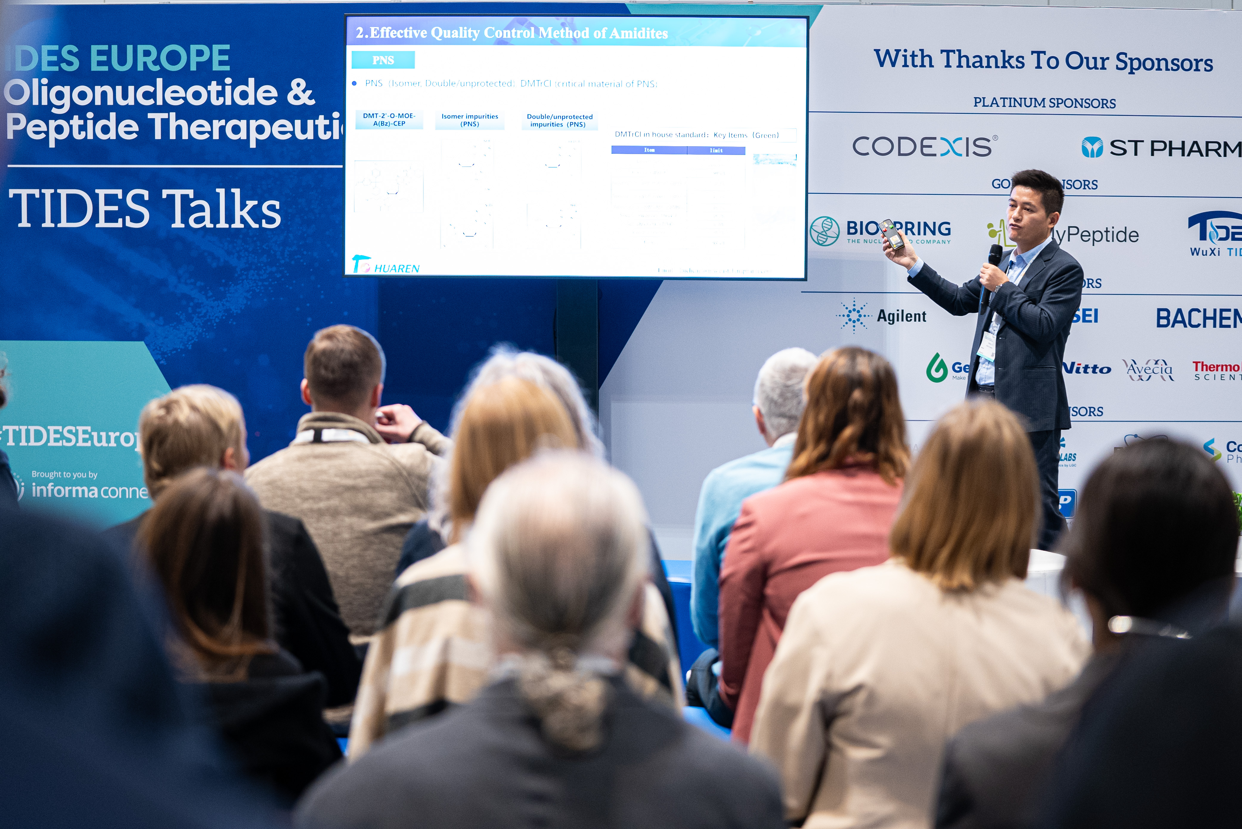 One speaker in a suit presents to an audience at TIDES EU pointing to a part of his presentation on screen.