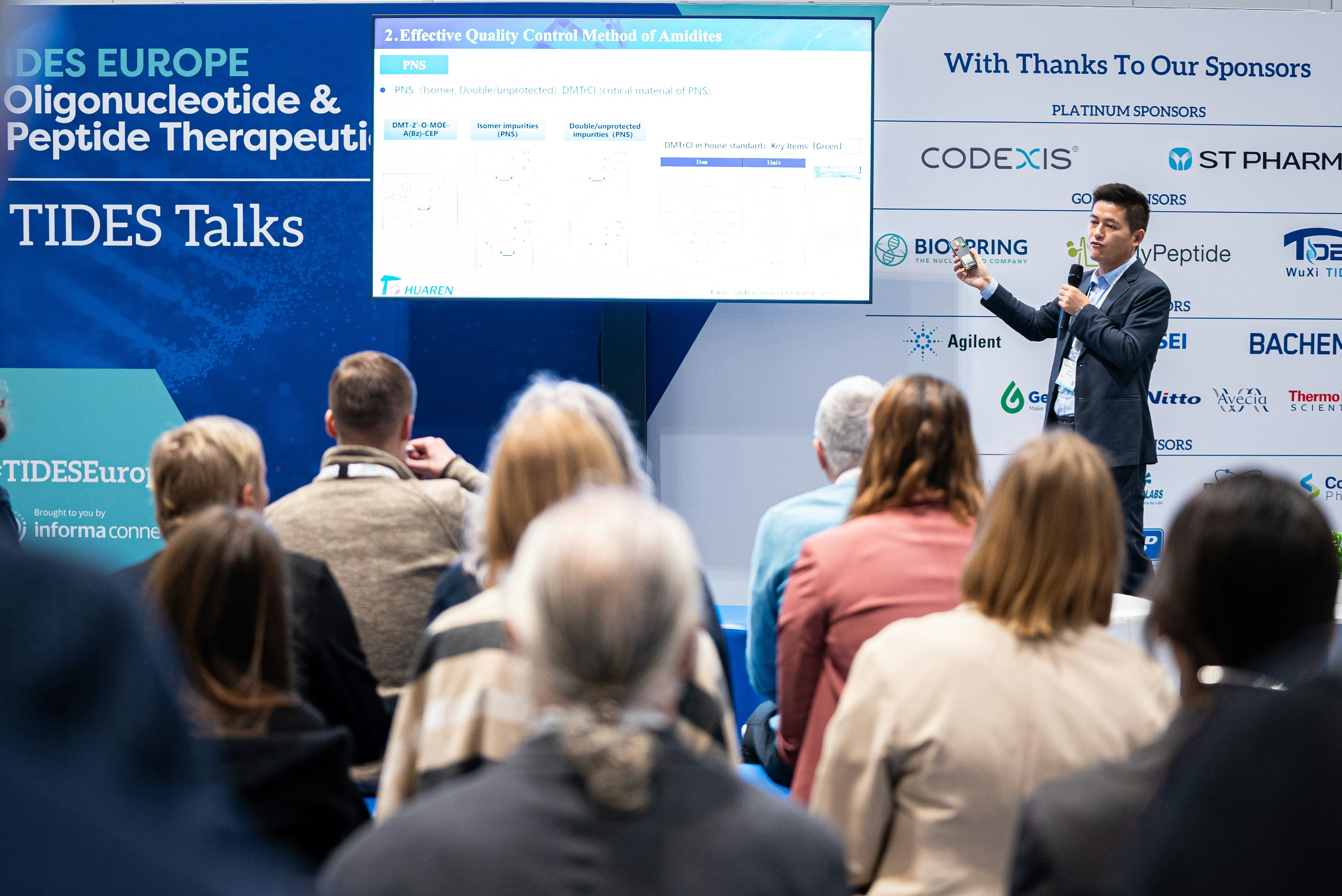 One speaker in a suit presents to an audience at TIDES EU pointing to a part of his presentation on screen.