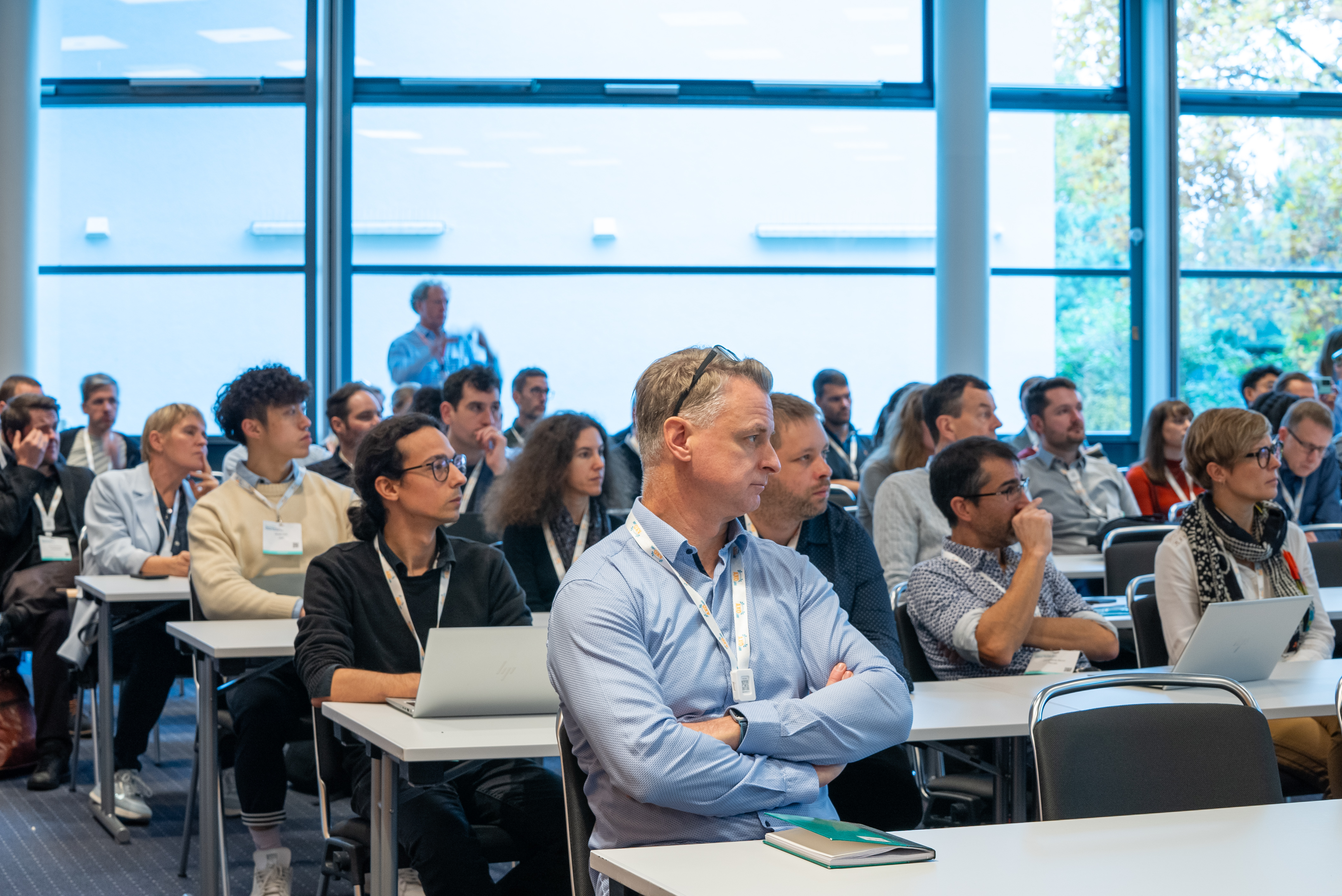 Attendees sitting in a conference room, one man in a blue shirt sits with his arms folded as he listens.