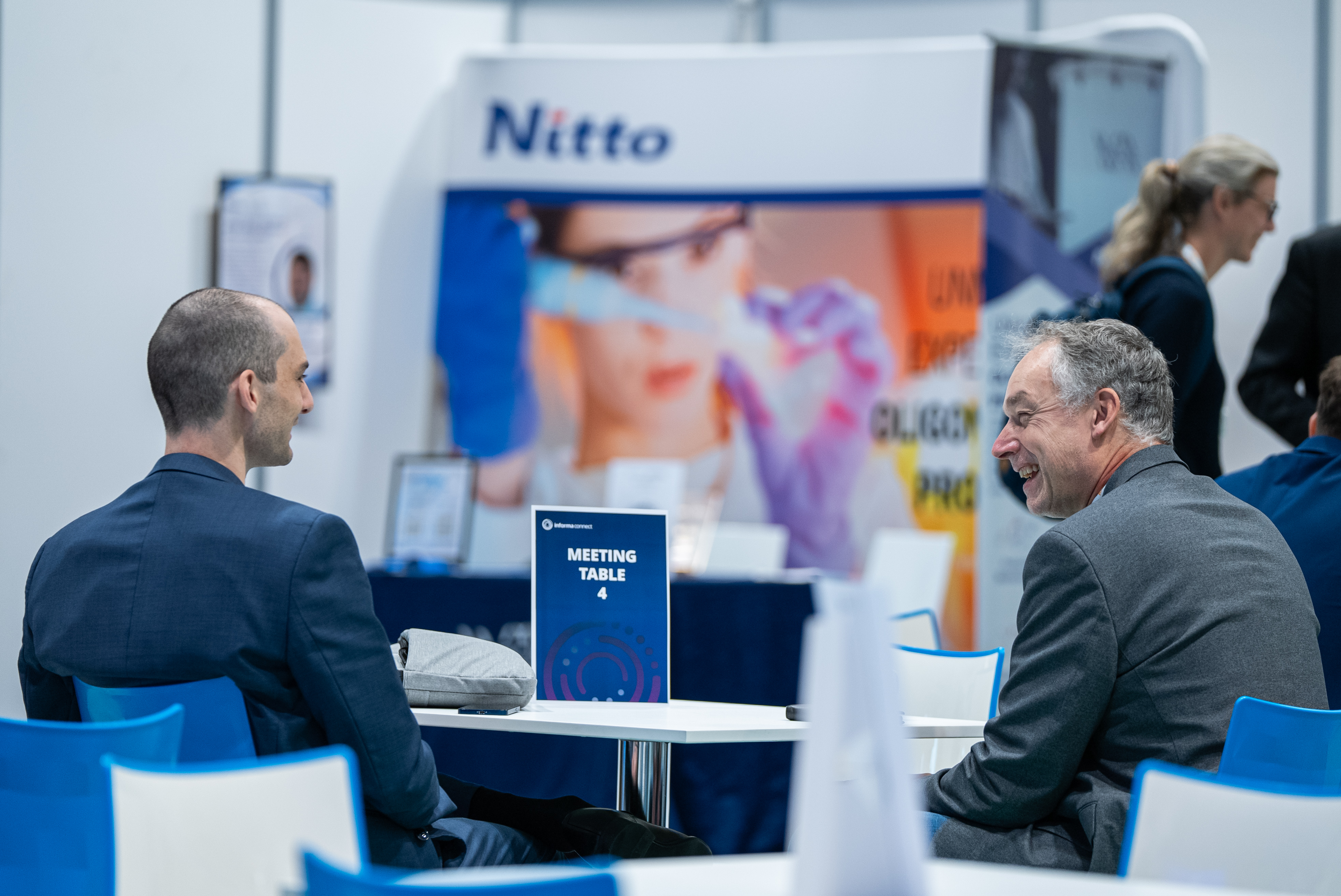 Two men in suits laugh with each other whilst sat at a meeting table.