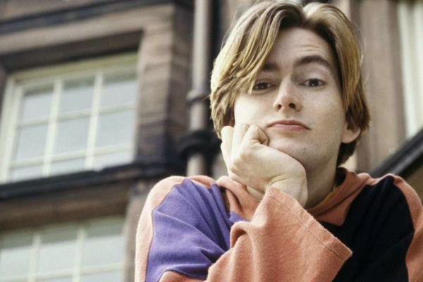 He is looking down with 90's style floppy blondish hair and he is leaning his chin on his hand