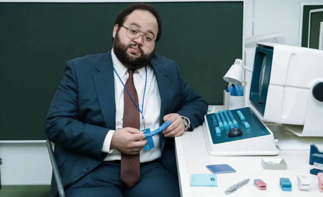 A man with dark hair and a bead sits in front of a 90s style computer. He is holding a blue finger trap, and surrounded by various coloured erasersr